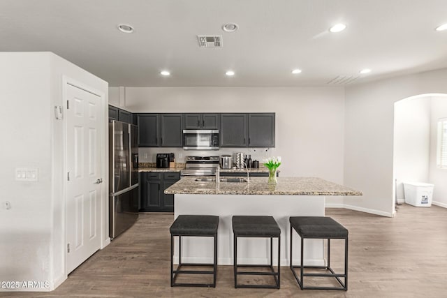 kitchen with appliances with stainless steel finishes, arched walkways, an island with sink, a kitchen bar, and visible vents
