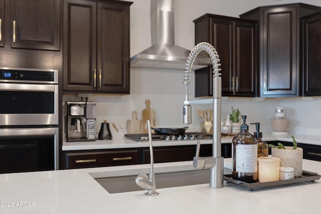 kitchen with dark brown cabinets, double oven, wall chimney exhaust hood, and light stone countertops