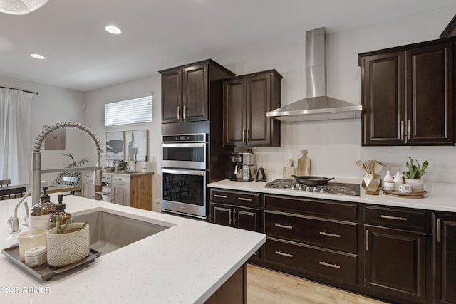 kitchen featuring dark brown cabinetry, appliances with stainless steel finishes, light wood-style floors, wall chimney exhaust hood, and a sink