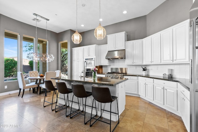 kitchen featuring appliances with stainless steel finishes, white cabinetry, range hood, decorative light fixtures, and a kitchen island with sink