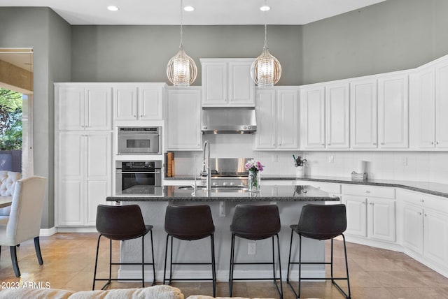 kitchen featuring hanging light fixtures, a kitchen island with sink, backsplash, white cabinetry, and ventilation hood