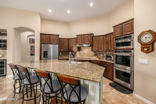 kitchen featuring decorative backsplash, appliances with stainless steel finishes, a kitchen island with sink, under cabinet range hood, and a sink