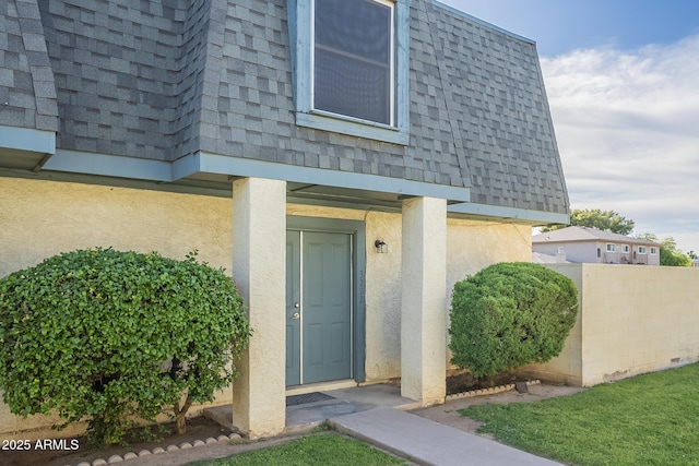 view of exterior entry with mansard roof, fence, stucco siding, and roof with shingles