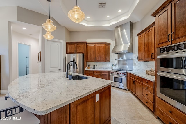 kitchen featuring pendant lighting, a kitchen island with sink, wall chimney exhaust hood, a tray ceiling, and stainless steel appliances