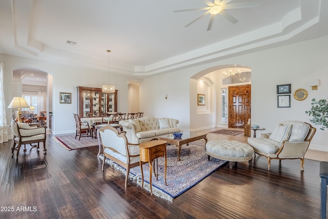 living room with ceiling fan with notable chandelier, a raised ceiling, and dark wood-type flooring