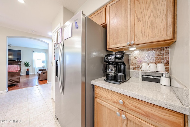 kitchen with light tile patterned floors, stainless steel fridge with ice dispenser, arched walkways, light brown cabinetry, and backsplash