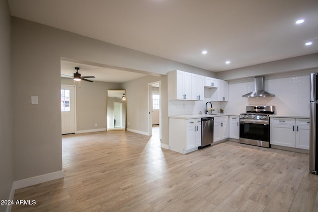 kitchen with stainless steel appliances, ceiling fan, wall chimney range hood, white cabinets, and light hardwood / wood-style floors