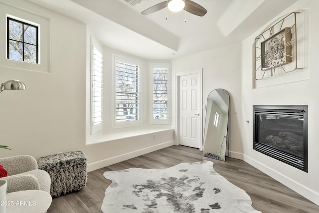 living area featuring a glass covered fireplace, ceiling fan, baseboards, and wood finished floors