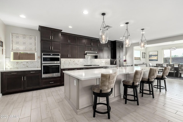 kitchen featuring decorative backsplash, a kitchen breakfast bar, light countertops, under cabinet range hood, and double oven