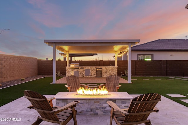 patio terrace at dusk featuring exterior kitchen, an outdoor fire pit, a fenced backyard, and a yard