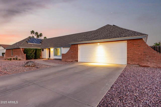 ranch-style house with a garage, brick siding, a shingled roof, driveway, and solar panels