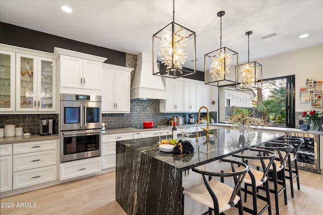 kitchen with a breakfast bar, stainless steel double oven, custom exhaust hood, dark stone counters, and a center island with sink