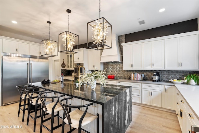 kitchen with a breakfast bar area, white cabinetry, dark stone countertops, an island with sink, and built in fridge