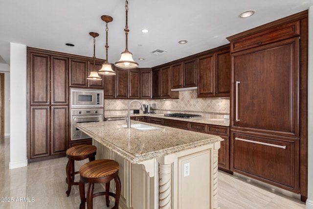 kitchen featuring visible vents, an island with sink, a sink, light stone counters, and stainless steel appliances