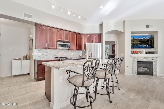 kitchen with a kitchen bar, a kitchen island with sink, decorative backsplash, stainless steel appliances, and light stone counters