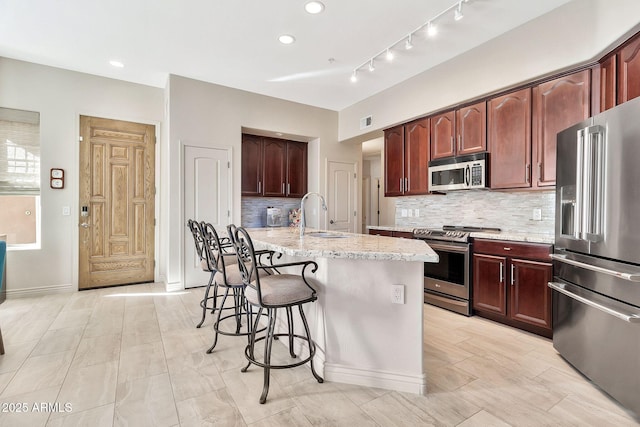 kitchen with stainless steel appliances, sink, backsplash, light stone counters, and a center island with sink