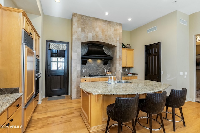 kitchen featuring light wood finished floors, visible vents, a kitchen island with sink, light stone countertops, and premium range hood