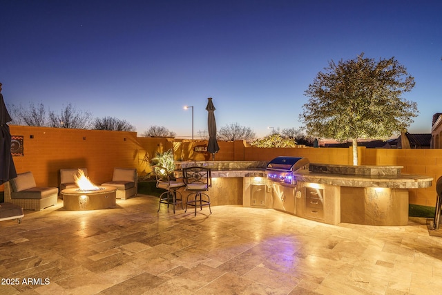 patio terrace at dusk with area for grilling, a fire pit, an outdoor bar, and exterior kitchen