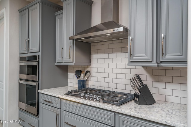 kitchen featuring wall chimney range hood, gray cabinetry, stainless steel appliances, light stone counters, and decorative backsplash