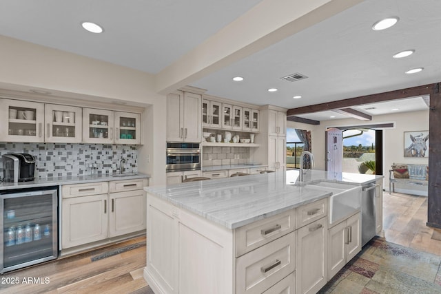 kitchen with a kitchen island with sink, wine cooler, glass insert cabinets, beamed ceiling, and backsplash