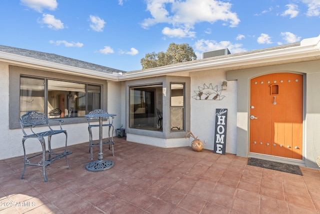 view of exterior entry with a shingled roof, a patio, and stucco siding