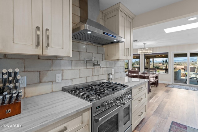 kitchen with light stone counters, wall chimney range hood, stainless steel stove, light wood finished floors, and decorative backsplash