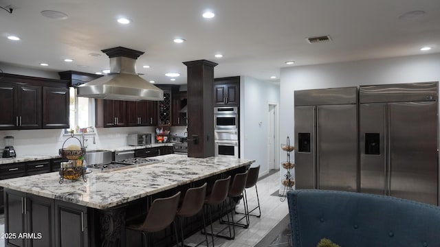 kitchen featuring island range hood, light stone counters, dark brown cabinetry, and appliances with stainless steel finishes