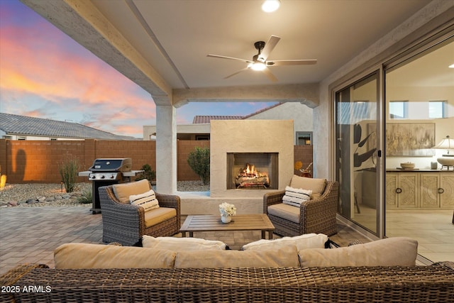 patio terrace at dusk with ceiling fan, an outdoor living space with a fireplace, fence, and grilling area