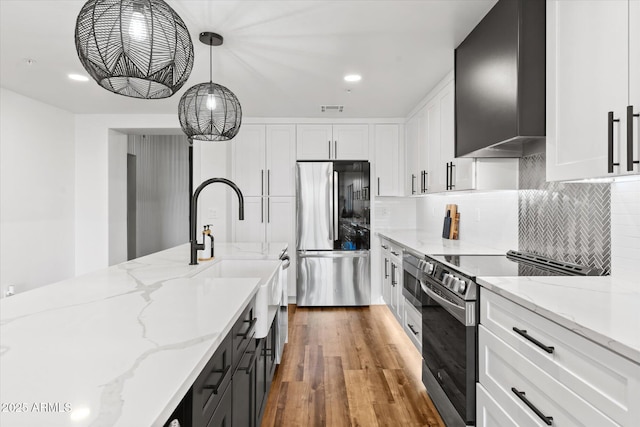 kitchen with white cabinets, wall chimney range hood, hanging light fixtures, light stone counters, and stainless steel appliances