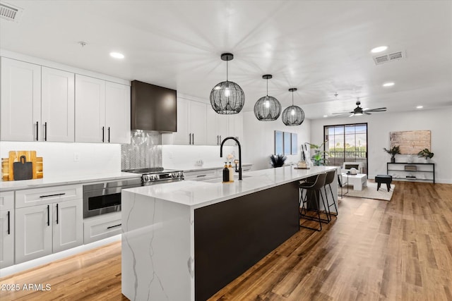 kitchen with a kitchen island with sink, wall chimney exhaust hood, ceiling fan, decorative light fixtures, and white cabinetry