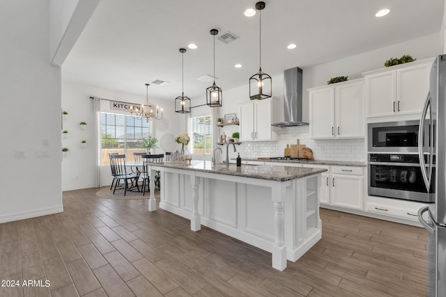 kitchen featuring appliances with stainless steel finishes, wall chimney exhaust hood, wood-type flooring, white cabinetry, and an island with sink