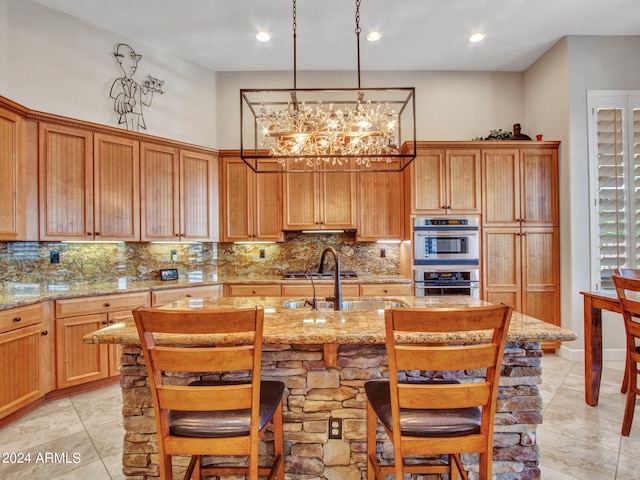 kitchen with appliances with stainless steel finishes, decorative backsplash, light stone countertops, an inviting chandelier, and a center island with sink