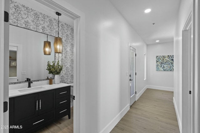 hallway with a sink, baseboards, wallpapered walls, light wood-style floors, and recessed lighting