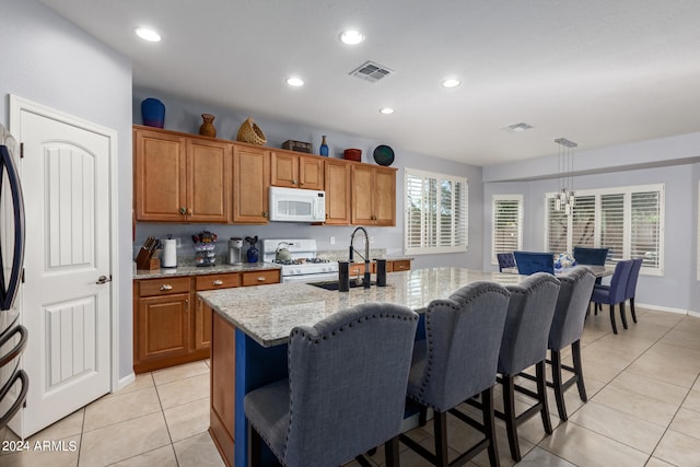 kitchen with light stone counters, a kitchen island with sink, sink, white appliances, and decorative light fixtures