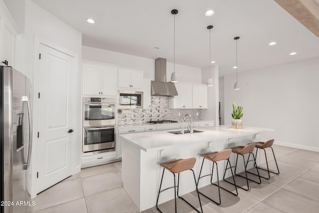 kitchen with white cabinetry, stainless steel appliances, wall chimney range hood, pendant lighting, and a kitchen island with sink