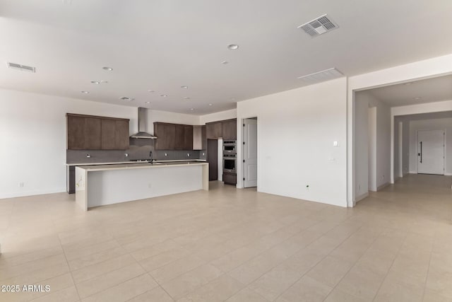 kitchen featuring open floor plan, wall chimney range hood, light countertops, a sink, and dark brown cabinets