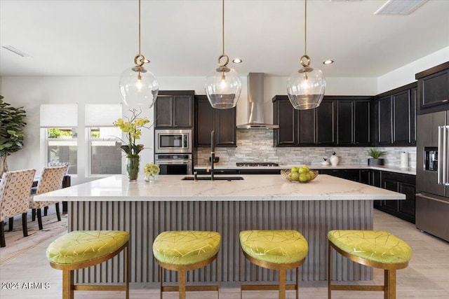 kitchen featuring wall chimney range hood, a kitchen island with sink, stainless steel appliances, and hanging light fixtures