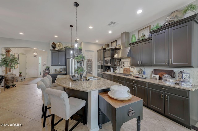 kitchen with arched walkways, stainless steel appliances, a sink, wall chimney range hood, and a kitchen bar