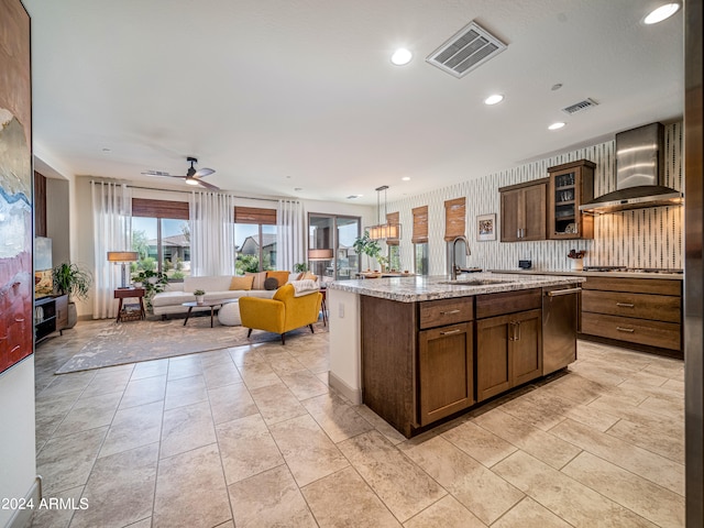 kitchen with a kitchen island with sink, sink, wall chimney exhaust hood, ceiling fan, and light stone counters