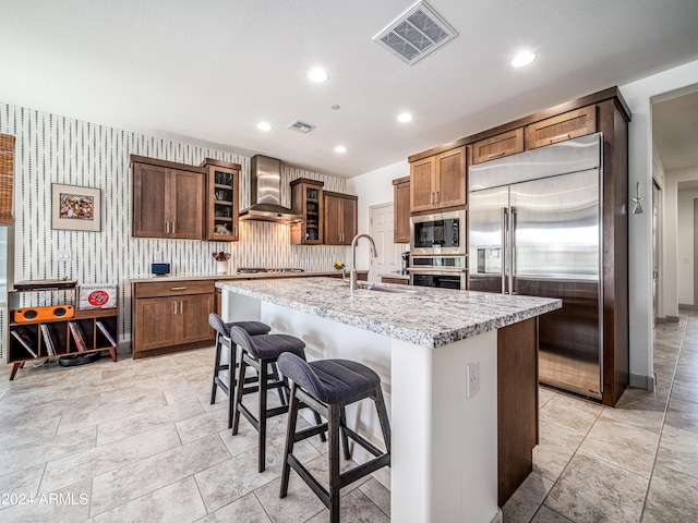 kitchen with a kitchen bar, sink, wall chimney range hood, built in appliances, and an island with sink