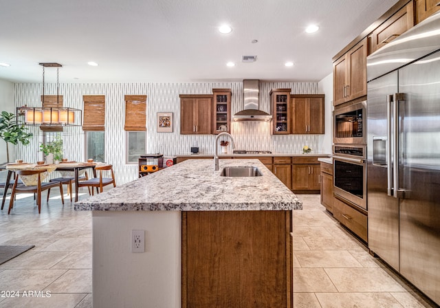 kitchen with wall chimney exhaust hood, a kitchen island with sink, sink, pendant lighting, and built in appliances