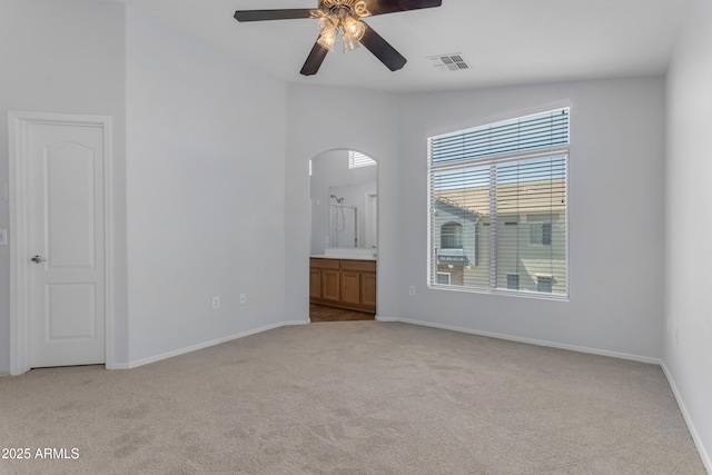 unfurnished bedroom with light carpet, arched walkways, baseboards, ensuite bathroom, and ceiling fan