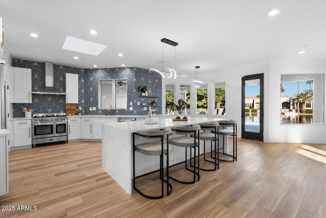 kitchen with decorative light fixtures, stainless steel appliances, white cabinets, an island with sink, and wall chimney exhaust hood