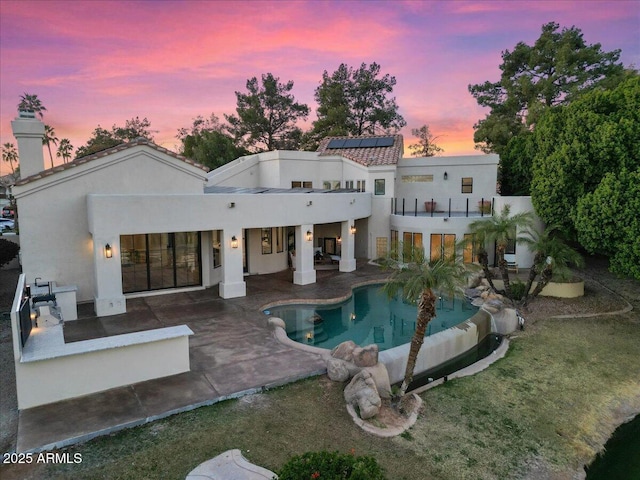 back of property at dusk featuring a balcony, an outdoor pool, roof mounted solar panels, stucco siding, and a patio area