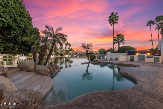 pool at dusk featuring a water view, a patio, grilling area, and an outdoor kitchen