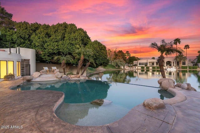 pool at dusk featuring a patio area, a water view, and an outdoor pool