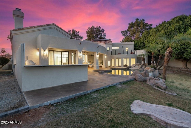 back of house at dusk featuring a yard, a chimney, a patio area, and stucco siding