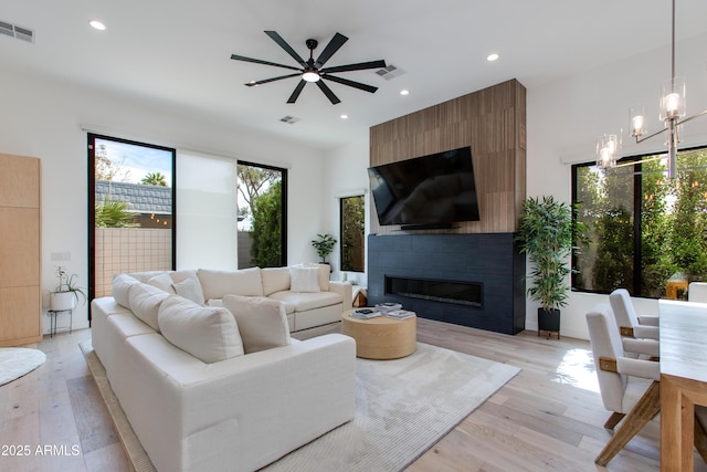 living area featuring a large fireplace, ceiling fan with notable chandelier, light wood finished floors, and visible vents