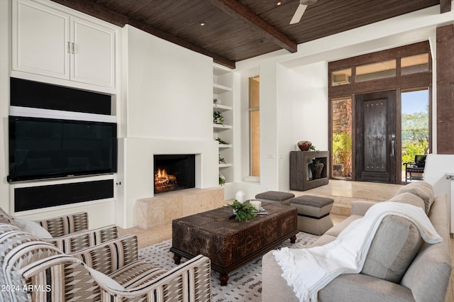 living area with built in shelves, beam ceiling, a ceiling fan, wooden ceiling, and a lit fireplace