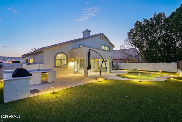 back house at dusk with exterior kitchen, a patio, and a lawn